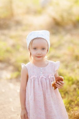 Hafif elbiseli ve kafasında eşarp olan bir kız tarlada bir keçi otlatıyor ve bir pasta, tarım, köyde bir çocuk yiyor.