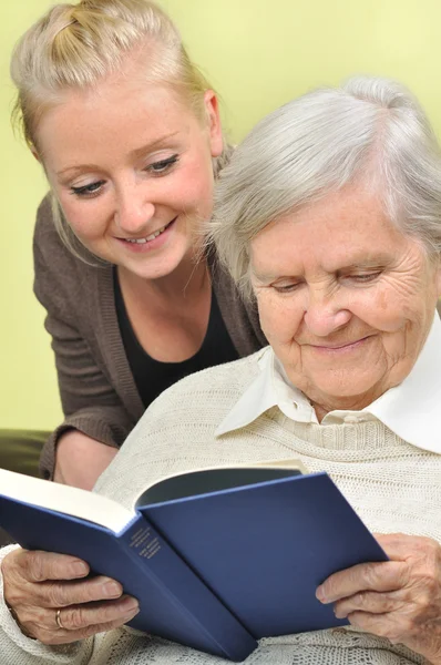 Senior woman with her caregiver in home reading book. — Stock Photo, Image