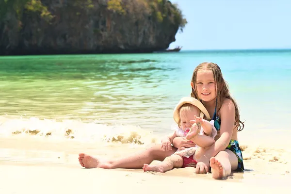 Två glada leende systrar sitter på en tropisk strand. — Stockfoto