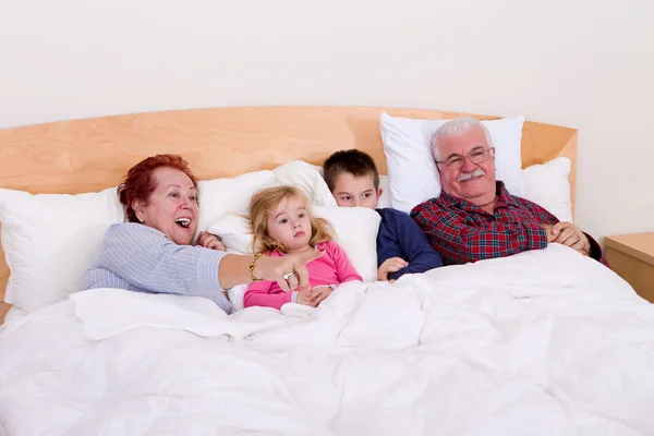 Großeltern, die mit ihren Enkeln im Bett fernsehen — Stockfoto