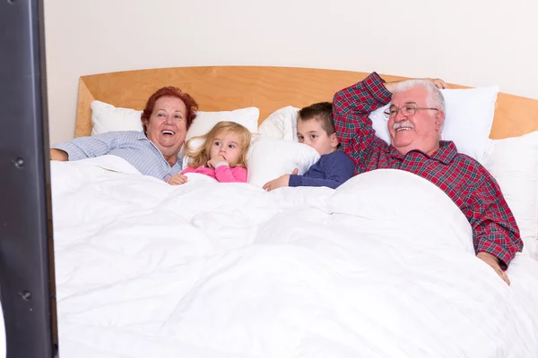 Großeltern, die mit ihren Enkeln im Bett fernsehen — Stockfoto