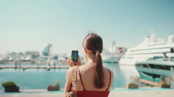 年轻女子用手机拍摄海港的照片和录像 女孩用社交网络和流媒体服务在智能手机上射击游艇和船只 背景色 — 图库视频影像