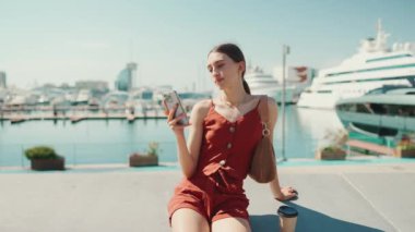 Limanda oturan güzel genç bir kadın bir cep telefonu kullanıyor. Kız arka plandaki yat ve gemilerdeki akıllı telefona bakıyor.