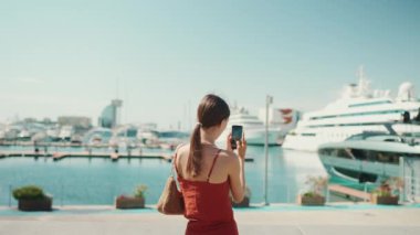 Şirin bir kadın, limanın videolarını cep telefonuyla çekiyor. Kız yat ve gemi vururken akıllı telefondan sosyal ağlar ve yayın hizmetleri kullanıyor.