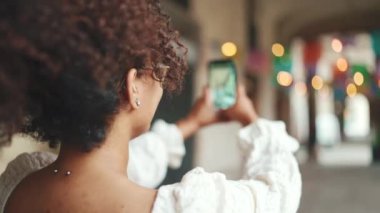 Gözlüklü genç kadın şehir merkezini gezerken akıllı telefondan fotoğraf çekiyor. Arkadan bak. Şehir arka planında cep telefonu kullanan olumlu kadın..