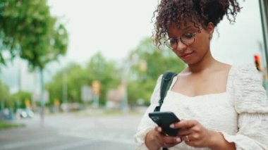 Sırt çantalı genç bir kadın caddede duran akıllı telefona bakıyor. Şehir arka planında cep telefonu kullanan olumlu kadın..