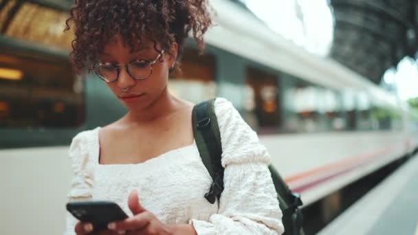 车站前 站着一个戴着眼镜 手持智能手机和背包的年轻女子的特写 城市背景下在室外使用手机的阳性妇女 — 图库视频影像