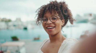 Gözlüklü genç bir kadın limanda dikiliyor ve akıllı telefondan selfie çekiyor. Şehir arka planında cep telefonu kullanan olumlu kadın..