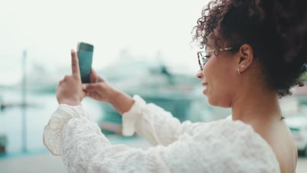 眼鏡をかけた若い女性が港に立ち スマートフォンで船を見て撮影する 都市部の屋外で携帯電話を使用して正の女性 — ストック動画