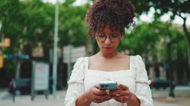 Gözlüklü genç bir kadın elinde akıllı telefonuyla ayakta duruyor ve etrafına bakıyor. Şehir arka planında cep telefonu kullanan kadın.