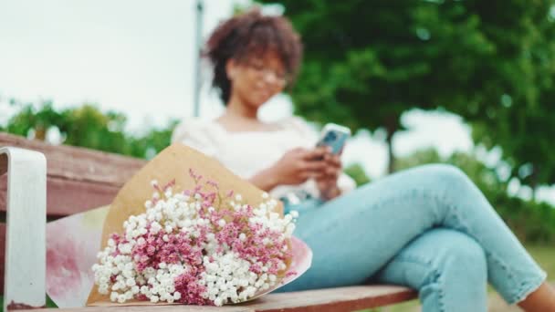 여자는 벤치에 스마트폰을 꽃다발을 있습니다 배경에서 핸드폰을 사용하는 긍정적 부드러운 — 비디오
