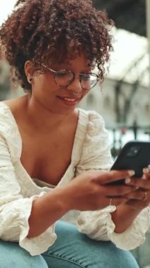 Gözlüklü genç bir kadının yakın plan portresi istasyonda oturuyor, akıllı telefona bakıyor ve gülümsüyor. Şehir arka planında cep telefonu kullanan pozitif kızın yakın plan fotoğrafı.