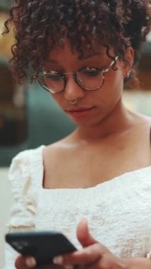 Gözlüklü, akıllı telefonlu ve sırt çantalı genç bir kadının istasyonda yakın plan portresi. Şehir arka planında cep telefonu kullanan pozitif kadının yakın plan fotoğrafı.