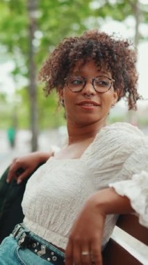 Gözlüklü genç bir kadının yakın plan portresi bir bankta oturuyor ve gülümsüyor ve etrafına bakıyor. Mutlu kızın yakın çekimi bir bankta otururken etrafına bakıyor.