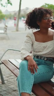 Gözlüklü genç kadının yakın plan portresi açık hava bankında oturuyor ve gülümsüyor ve akıllı telefonda konuşuyor. Şehir arka planında cep telefonu kullanan pozitif kadının yakın plan fotoğrafı.