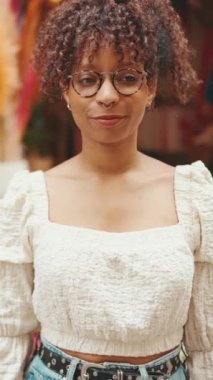 Gözlüklü genç bir kadının yakın plan portresi bir çiçekçinin yanında duruyor ve kameraya bakıyor ve gülümsüyor. Kamerada poz veren kız