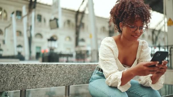 Mujer Joven Gafas Sienta Estación Mira Teléfono Inteligente Sonríe Mujer — Vídeos de Stock