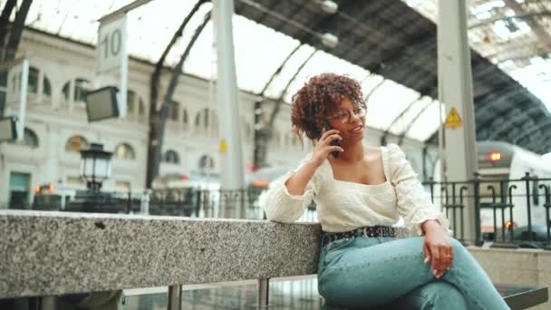 一个戴眼镜的年轻女人坐在车站 在电话里聊天 城市背景下在户外使用手机的积极女性 — 图库视频影像