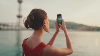 Yakın plan, kız setin üzerinde duruyor ve cep telefonuyla güneşin doğuşunun fotoğraflarını çekiyor. Genç bir kadın sabahları akıllı telefondan denize ateş ediyor..