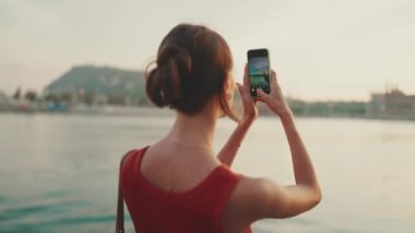 Yakın plan, kız setin üzerinde duruyor ve cep telefonuyla güneşin doğuşunun fotoğraflarını çekiyor. Genç bir kadın, sabahları akıllı telefonuyla denizi vuruyor. Arka plan