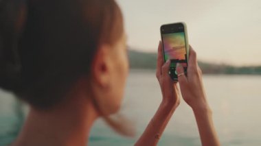 Yakın plan, kız setin üzerinde duruyor ve cep telefonuyla güneşin doğuşunun fotoğraflarını çekiyor. Genç bir kadın, sabahları akıllı telefonuyla denizi vuruyor. Arka plan