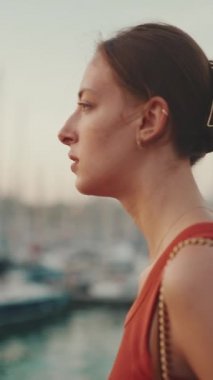 Yakından, gezinti güvertesi boyunca kız limandaki yatlara bakıyor ve saçlarını düzleştiriyor. Genç bir kadının yakın plan portresi sabah vakti deniz boyunca yürür..