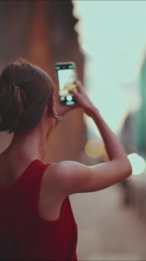 Yakın çekim, kız şehir merkezindeki cep telefonunun resimlerini çekiyor. Genç bir kadının yakınlaşması, şehir manzarasını akşam saatlerinde akıllı telefondan çeker. Arka plan