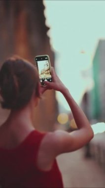 Yakın çekim, kız şehir merkezindeki cep telefonunun resimlerini çekiyor. Genç bir kadının yakınlaşması, şehir manzarasını akşam saatlerinde akıllı telefondan çeker. Arka plan