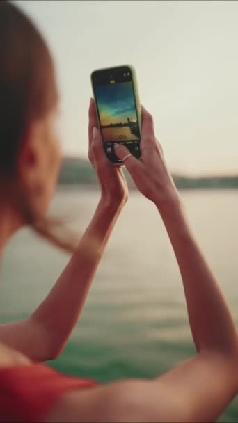 垂直方向のビデオ クローズアップ 少女は堤防の上に立って 携帯電話で日の出の写真を撮る 若い女性の閉鎖は 午前中にスマートフォンで海を撮影します バックビュー — ストック動画