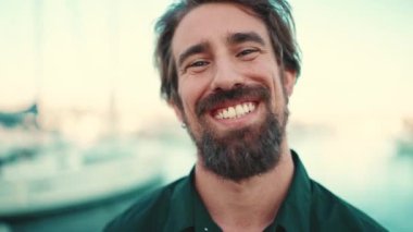 Deniz kenarında, yat arka planında sakallı, gülümseyen bir adamın yakın plan portresi. Kameraya bakan genç ve mutlu hipster erkeğin önden yakın çekimi.
