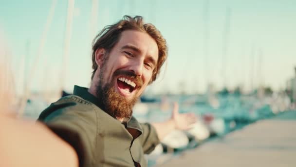 一个满脸笑容的男人 留着胡子 在堤岸上 在游艇的背景上聊天 他的服装肖像 用手机对快乐的年轻男性嬉皮士进行前部特写 — 图库视频影像