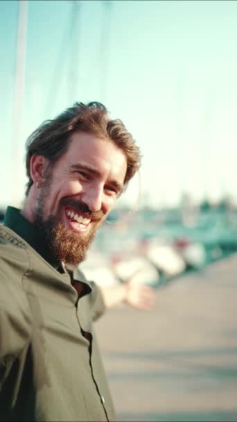 垂直录像 一个满脸笑容的男人 留着胡子 在堤岸上 在游艇的背景上聊天的合影 用手机对快乐的年轻男性嬉皮士进行前部特写 — 图库视频影像