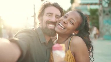 Irklar arası gülümseyen, dondurma yiyen ve şehir geçmişinde selfie çeken bir çiftin yakın plan portresi. Yakın plan, kadın ve erkek dondurma tadıp cep telefonuyla görüntülü sohbet ediyorlar. Arkaplan