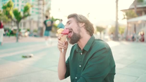 在城市背景上 一个留着胡子 手里拿着冰激凌的笑脸男子的特写 在一个温暖的阳光灿烂的日子里 一个快乐的年轻嬉皮士男子正在做冰淇淋的前置特写 — 图库视频影像