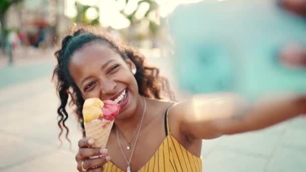 一个微笑的年轻女子 留着长长的卷发 手里拿着冰淇淋 在城市背景的手机上打一个视频电话 一个快乐女孩用智能手机进行交流的特写镜头 — 图库视频影像