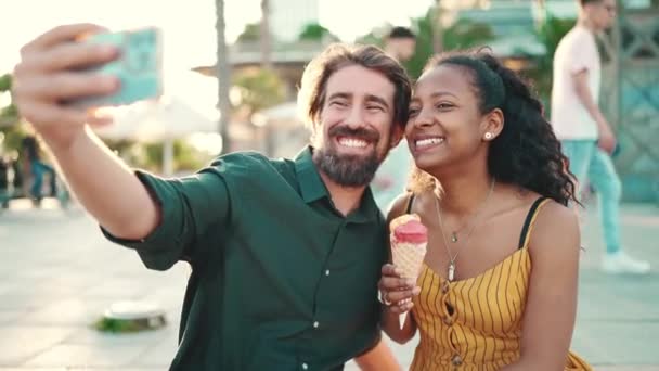 笑着的跨种族夫妇一起吃冰淇淋 并在城市背景下自拍的服装 一个男人和女人用手机品尝冰淇淋和视频聊天的特写镜头 — 图库视频影像