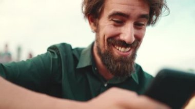 Arka planda sakallı, gülümseyen bir adamın yakın plan portresi. Cep telefonu kullanan genç ve mutlu erkek hippilerin önden yakın çekimi.