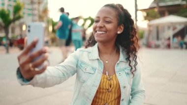 Kentin arka planında cep telefonu kullanan uzun kıvırcık saçlı genç bir kadının yakın plan portresi. Kız fotoğraf çekerken, akıllı telefondan video görüşmesi yaparken.