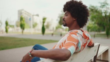 Parktaki bankta rahatça oturan, gömlek giyen Afro-Amerikalı genç adam başka tarafa bakıyor. Yaşam tarzı konsepti. Yavaş çekim