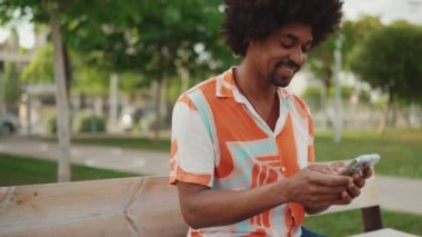 Parktaki bankta oturan Afro-Amerikan gömlekli genç adam akıllı telefonunu kullanıyor. Gülümseyen Afrikalı Amerikalı adam sosyal ağ hizmetlerinde cep telefonuyla sohbet ediyor.