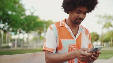Parktaki bankta oturan Afro-Amerikan gömlekli genç adam akıllı telefonunu kullanıyor. Sosyal ağ hizmetlerinde iletişim kuran adam. Yaşam tarzı konsepti. Yavaş çekim
