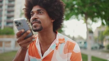 Parktaki bankta oturan Afro-Amerikan gömlekli genç adam akıllı telefonunu kullanıyor. Gülümseyen adam cep telefonundan sesli mesaj gönderiyor. Yaşam tarzı konsepti. Yavaş çekim