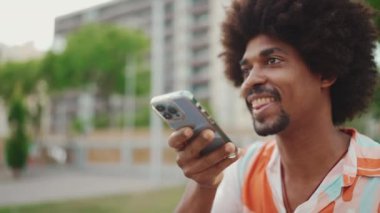 Parktaki bankta akıllı telefonuyla oturan, gömlek giyen Afro-Amerikan bir adamın yakın plan portresi. Gülümseyen adam cep telefonundan sesli mesaj gönderiyor. Kamera adamın etrafında dönüyor.