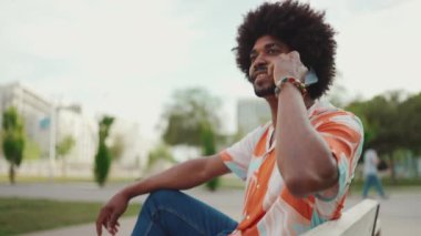 Parktaki bankta oturan Afro-Amerikan gömlekli genç adam akıllı telefonunu kullanıyor. Cep telefonuyla konuşan gülen adam. Kamera adamın etrafında dönüyor. Yavaş çekim
