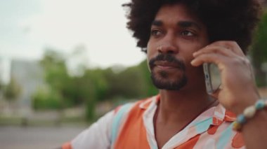 Parktaki bankta oturan Afro-Amerikan gömlekli genç adamın akıllı telefonunu kullanarak yakın plan portresi. Adam cep telefonuyla konuşuyor ve saçını düzeltiyor. Yaşam tarzı konsepti. Yavaş çekim