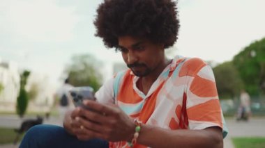 Parktaki bankta oturan Afro-Amerikan gömlekli genç adam akıllı telefonunu kullanıyor. Gülümseyen Afro-Amerikan bir adamın cep telefonundaki fotoğraf videosuna yakından bakması. Yaşam tarzı kavramı