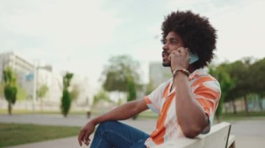 Parktaki bankta oturan Afro-Amerikan gömlekli genç adam akıllı telefonunu kullanıyor. Cep telefonuyla konuşan ve başını sallayan bir adam. Yaşam tarzı konsepti. Yavaş çekim