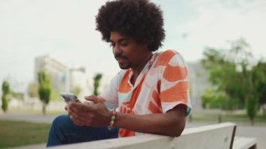 Parktaki bankta akıllı telefonuyla oturan, gömlek giyen Afro-Amerikan bir adamın yakın plan portresi. Yaşam tarzı konsepti. Yavaş çekim