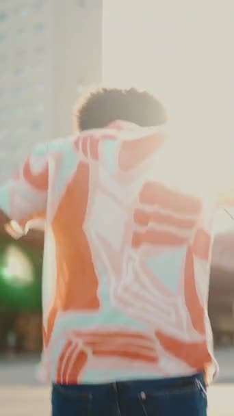 Verical Video 明るいアフリカ系アメリカ人の若者が ヘッドフォンで音楽を聴いたり 都会の街の背景で踊ったりするのを間近で見ることができます ライフスタイルの概念 バックライトスローモーション — ストック動画