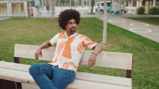 坐在公园的长椅上 穿着衬衫的年轻非洲裔美国人的特写镜头放松了下来 他们把目光移开 触摸着他的下巴 生活方式的概念 慢动作 — 图库视频影像
